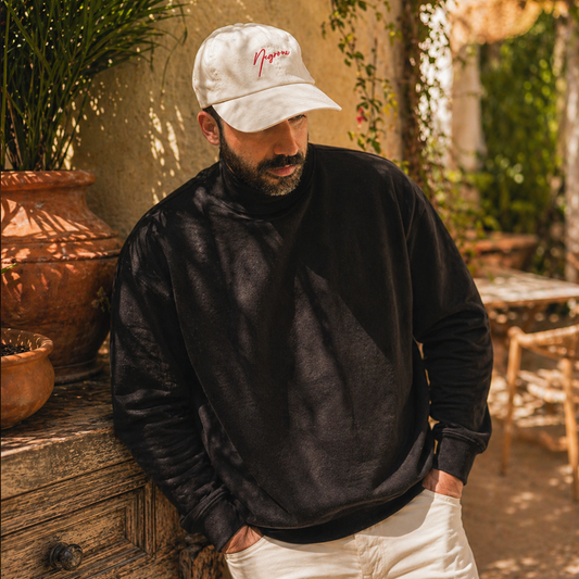 Man wearing a black sweatshirt and white cap in an outdoor setting with plants and furniture.